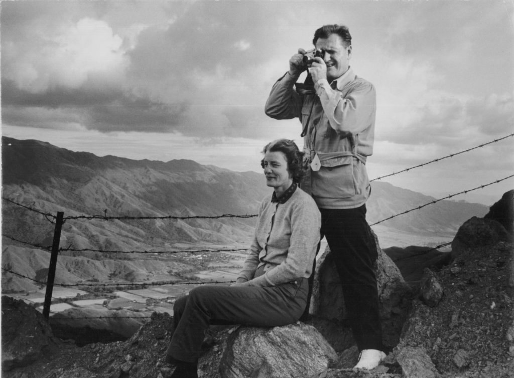 A black-and-white image of Nelson Rockefeller (who appears to be standing above a seated Tod) at their ranch in Venezuela. The mountains can be seen in the distance behind a wired fence