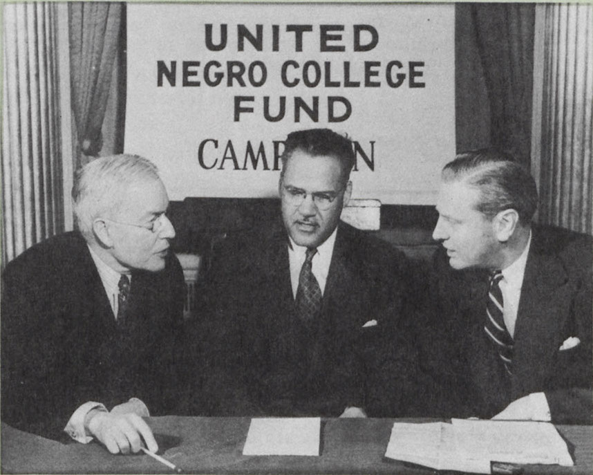 A black-and-white image of John D. Rockefeller and two UNCF officials, all dressed in formal attire. All are Caucasian and male. In the background hangs a sign that reads "United Negro College Fund Campaign."