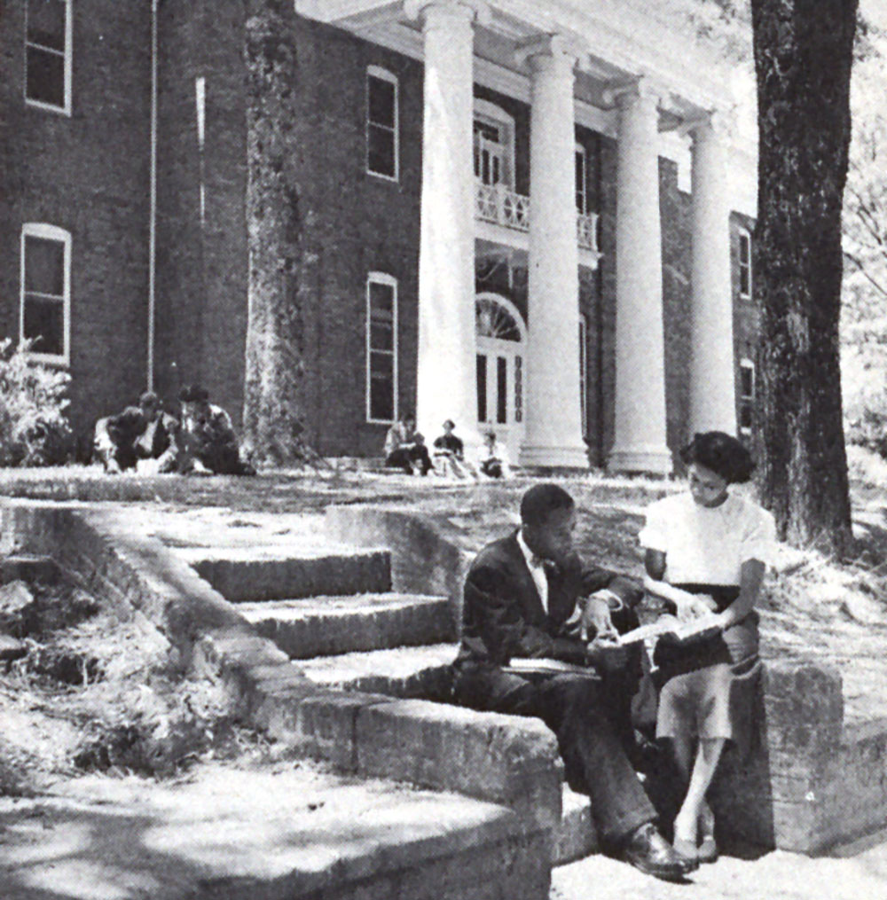 A black-and-white image of two African American students (one male, and one female), reading a book together. They are sitting on the steps of a university campus.