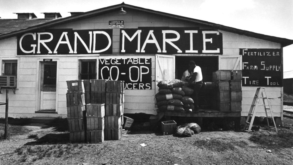 Black and white photo of "Grand Marie", a vegetable Co-Op. Crates are stacked in front of the signage, as two men are standing in front of two doors next to other farming supplies.