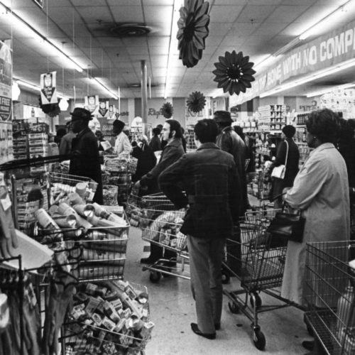 Black and white photo of the first grocery store in the area. Featured are long lines of individual's waiting in line to purchase goods.