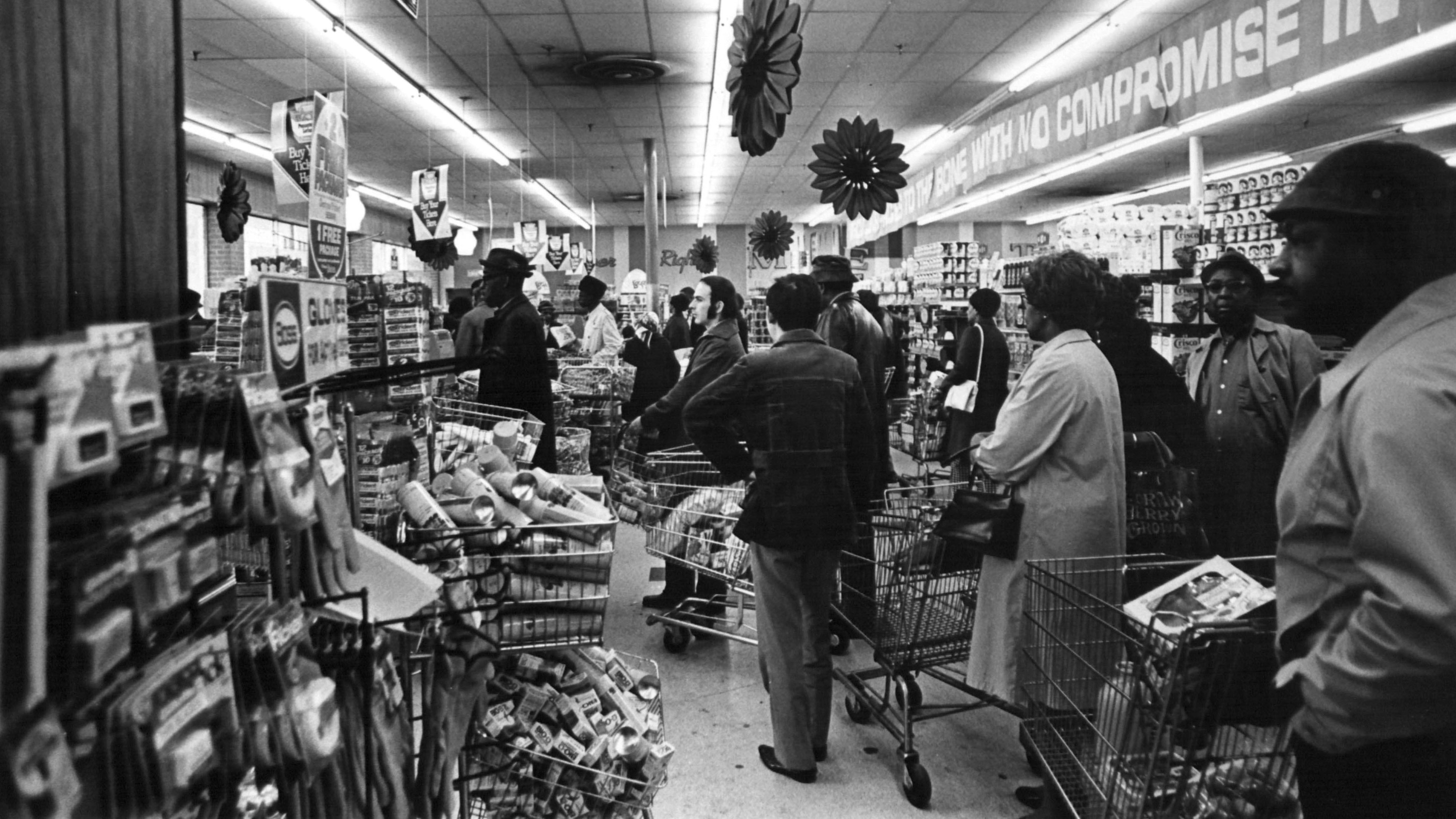 Black and white photo of the first grocery store in the area. Featured are long lines of individual's waiting in line to purchase goods.