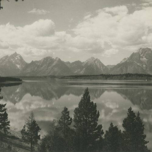 A sepia- toned landscape photograph of the Grand Teton National Park. The mountain range are reflected onto the lake while the trees frame the shot.