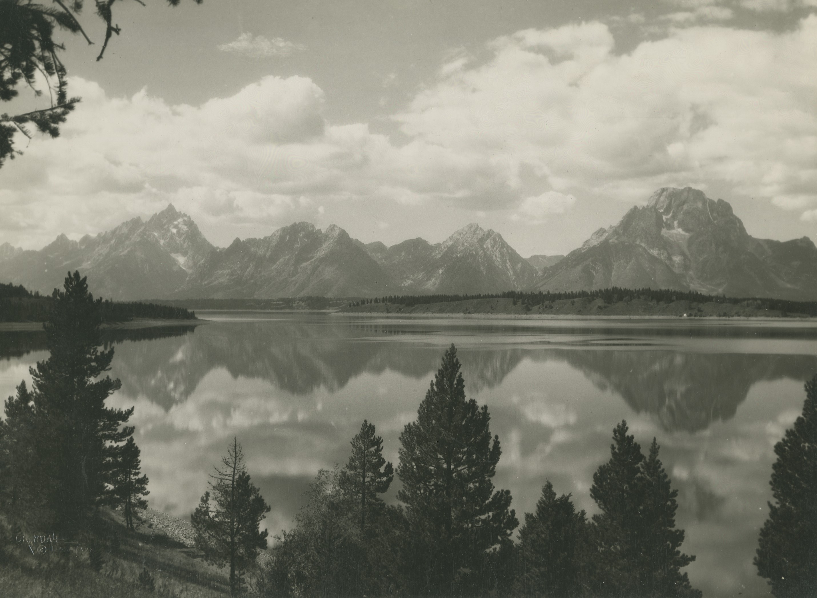 A sepia- toned landscape photograph of the Grand Teton National Park. The mountain range are reflected onto the lake while the trees frame the shot.