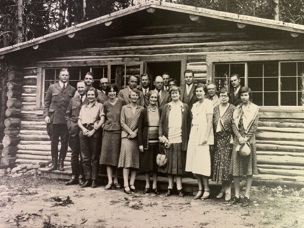 A sepia-toned photograph (dated 1930) of the "Rockefeller Travelling Party", taken in 1930. In the image are 12 men (in the back row) and 8 women (in the front row), all Caucasian. They are standing in front of a log cabin, "Old Elbo Ranch" located in Jackson Hole.