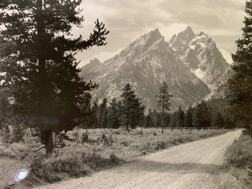 A sepia-toned image of a dirt road bending towards the left, while the Teton Mountain Range can be seen in the distance