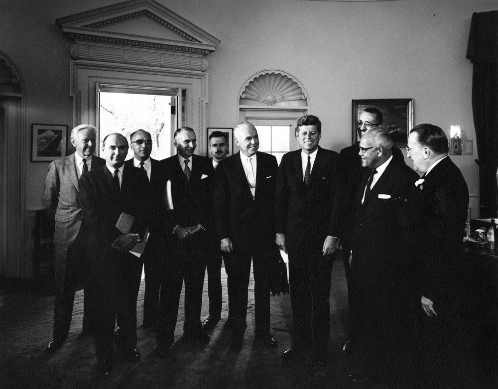 A black-and-white image of John F. Kennedy, surrounded by 9 members of the UNCF who are all directly facing him with enthusiastic expressions. They appear to be in the Oval office of the White House.