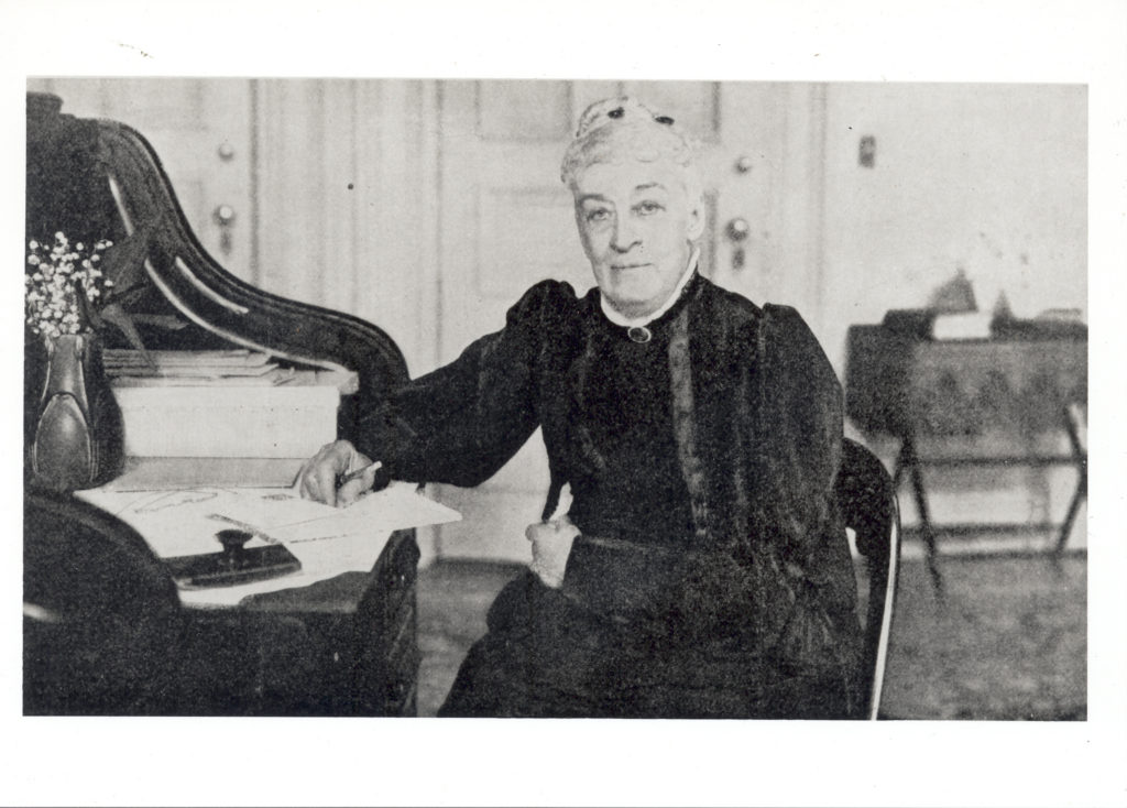 In this image, Margaret Sage, founder of the Sage Foundation is seated at a desk, with pen and paper in hand. She is dressed in her formal attire.