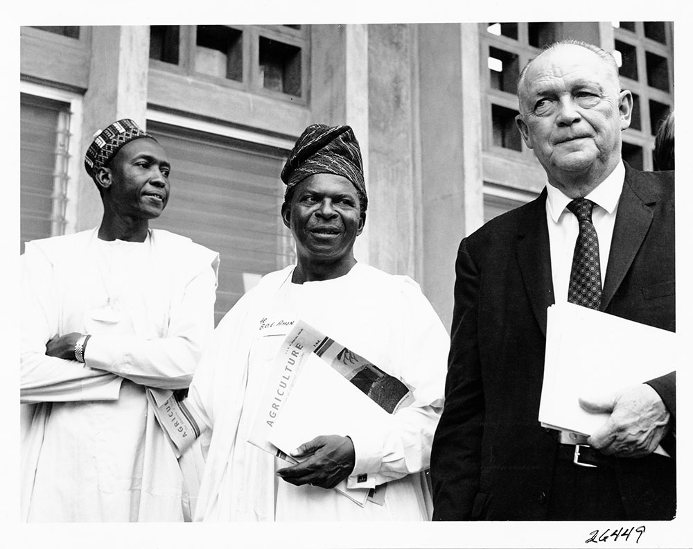 Rockefeller Foundation president, George Harrar next to two unnamed Nigerian officials. Harrar, holding a closed folder of documents appears stern as the two officials gaze at his skeptically. They are holding newspapers that read "agriculture" in bold lettering.
