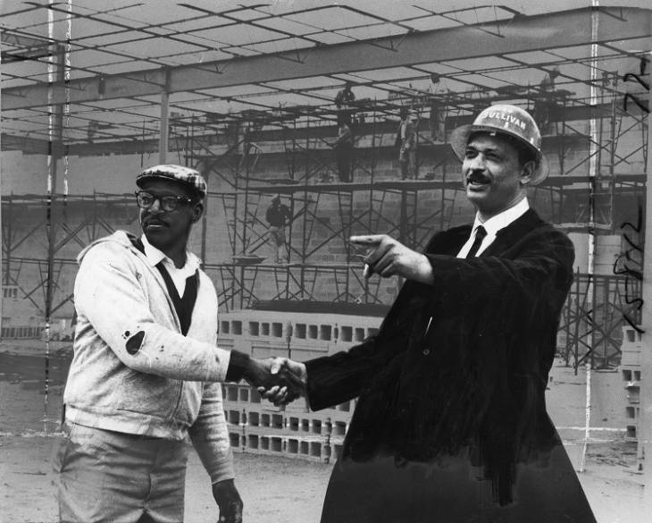 Two African- American men shaking hands, while the man on the right points his finger to the left. Behind them are scaffolding and construction workers