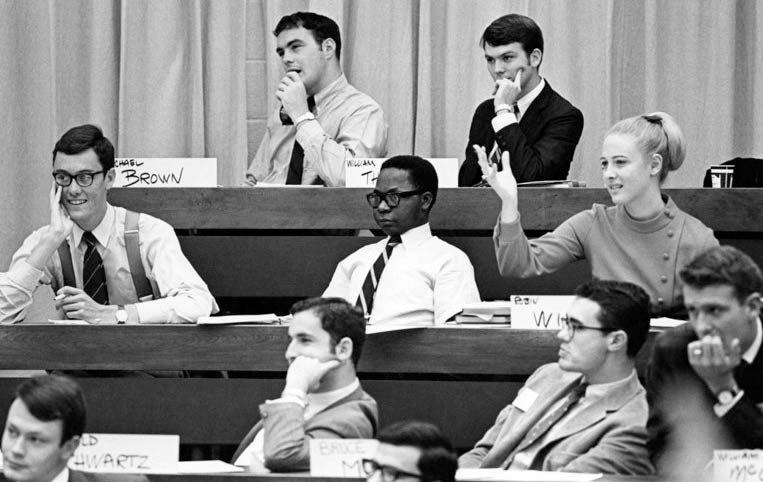 Nine students listen to a lecture in a lecture hall. A female student raises her hand.