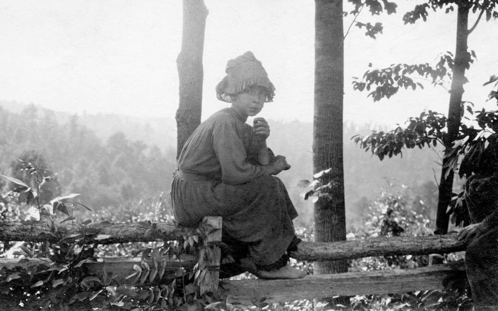 Young girl sitting on top of a fence with a hat and no shoes on in a wooded area.