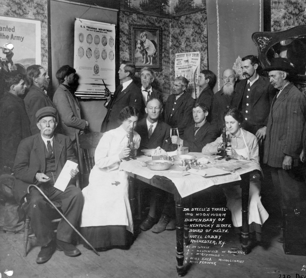 Dr. Steele's Traveling Hookworm Dispensary of Kentucky State Board of Health. Group of 15 looking at a poster distributed by the U.S Treasury Department, with another group seated at the table with microscopes and other scientific equipment.