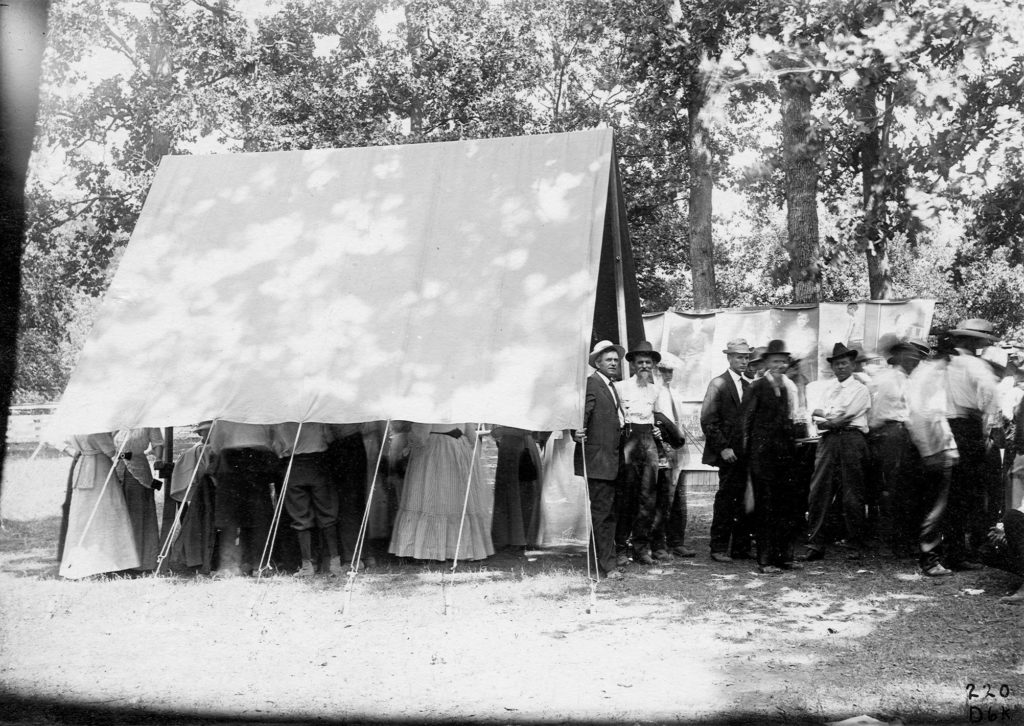 Black and white image of a group passing under a tent to view large prints of patients infected with hookworm.
