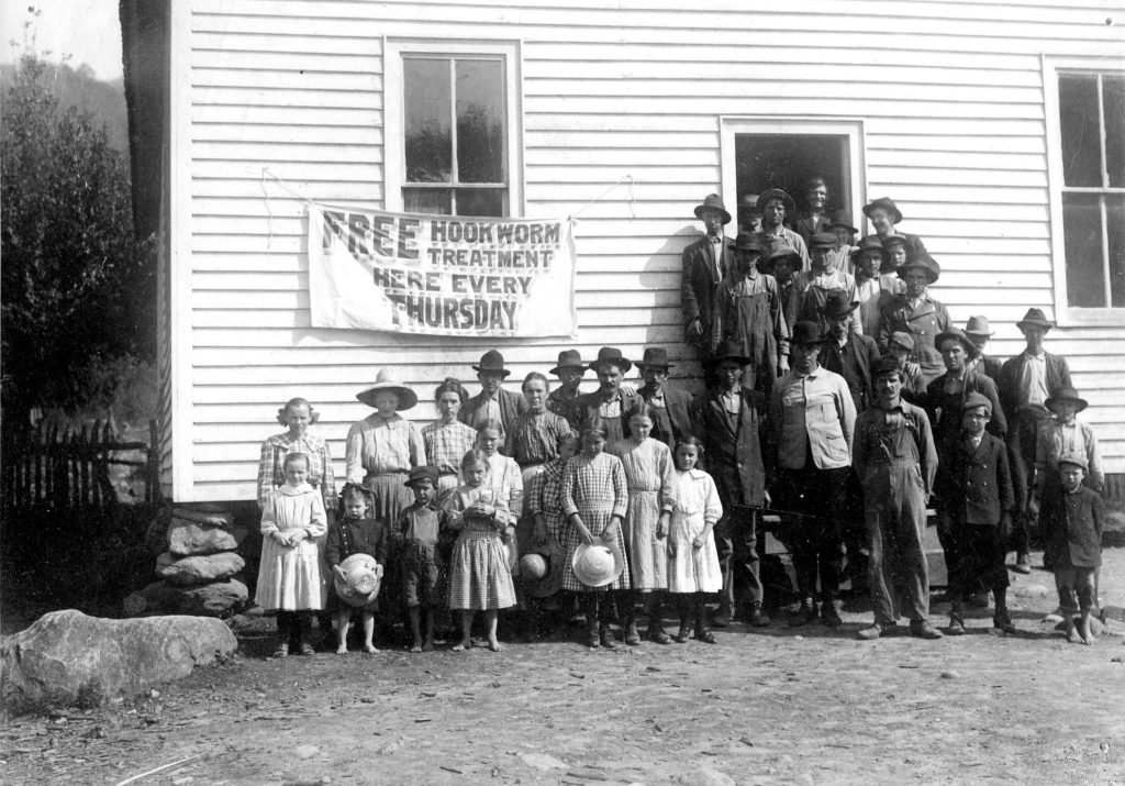 Black and white image of vistors to the Free Hookwork Treatment Center. Group of indivduals stand on both the stiarwell and underneath the banne that reads "Free Hookworm Treatment Here Evey Thursday"