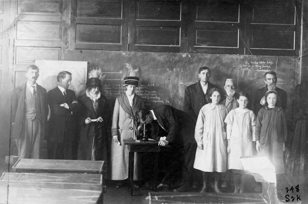 Group of eleven people standing in the front of a classroom as Professor T.E.R Davis looks into a microscope.