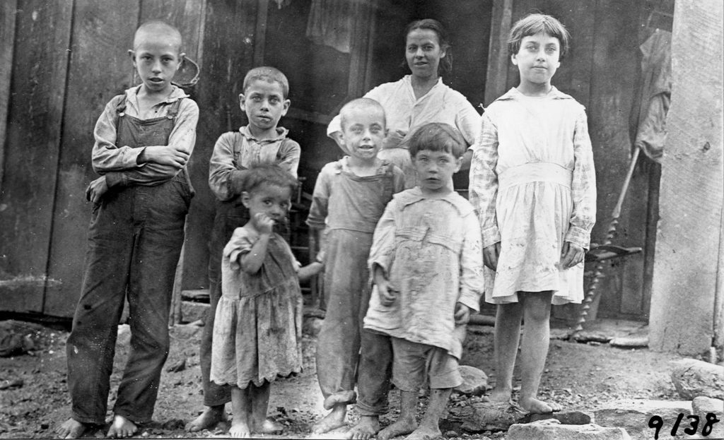 Six children standing with their mother outside of their home. Children are standing in moist soil without shoes.