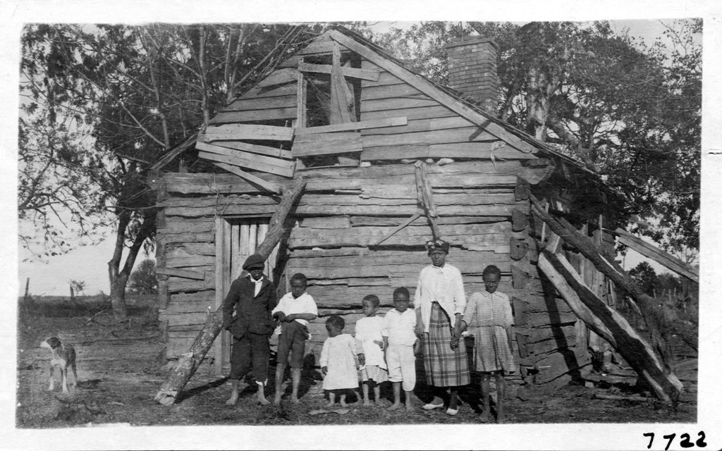 Seven children standing in front of their home, covered in wooden singles and being held up by tree logs. All but one person in the photo is wearing shoes.