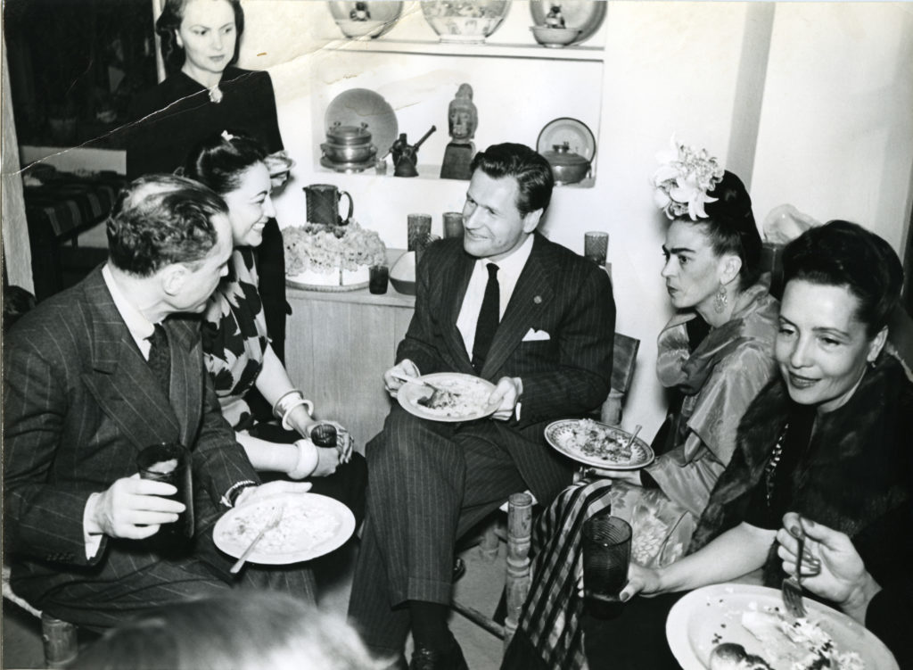 black-and-white image of 6 people (including Nelson Rockefeller and Frida Kahlo) gathered for a meal in Mexico circa 1943