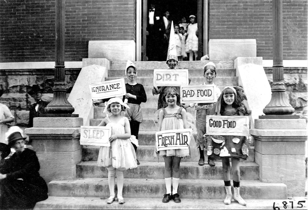 Children hold signs with words relating to hygiene. The signs read, in clockwise order, "ignorance, dirt, bad food, good food, fresh air, sleep." The work was funded by the Rockefeller Sanitary Commission for the Eradication of Hookworm.