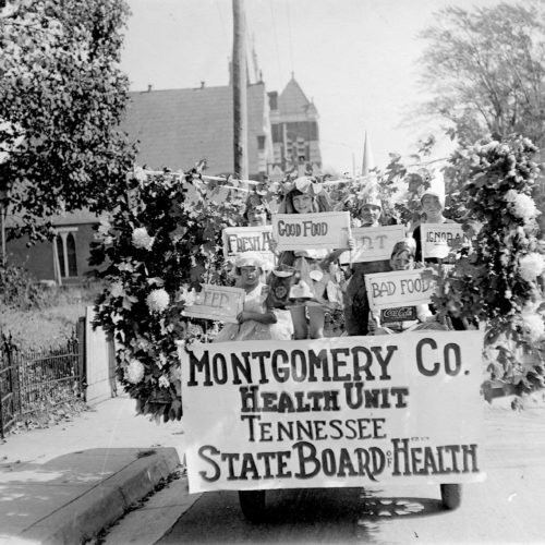 Black and white image of a group of children on a cart that is decorated with them holding signs reading "sleep", "fresh air", "good food" "dirt" and "ignorance".
