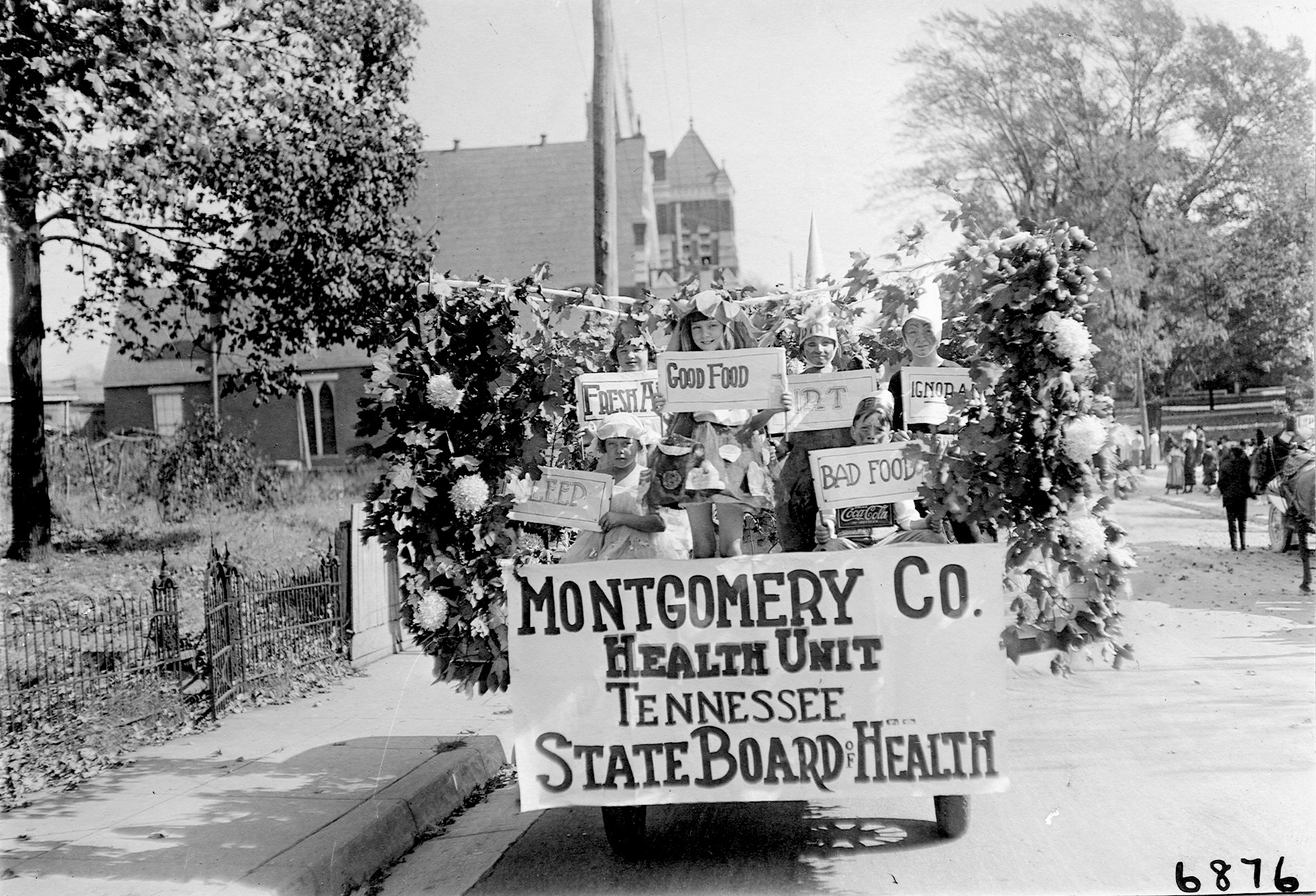 Black and white image of a group of children on a cart that is decorated with them holding signs reading "sleep", "fresh air", "good food" "dirt" and "ignorance".