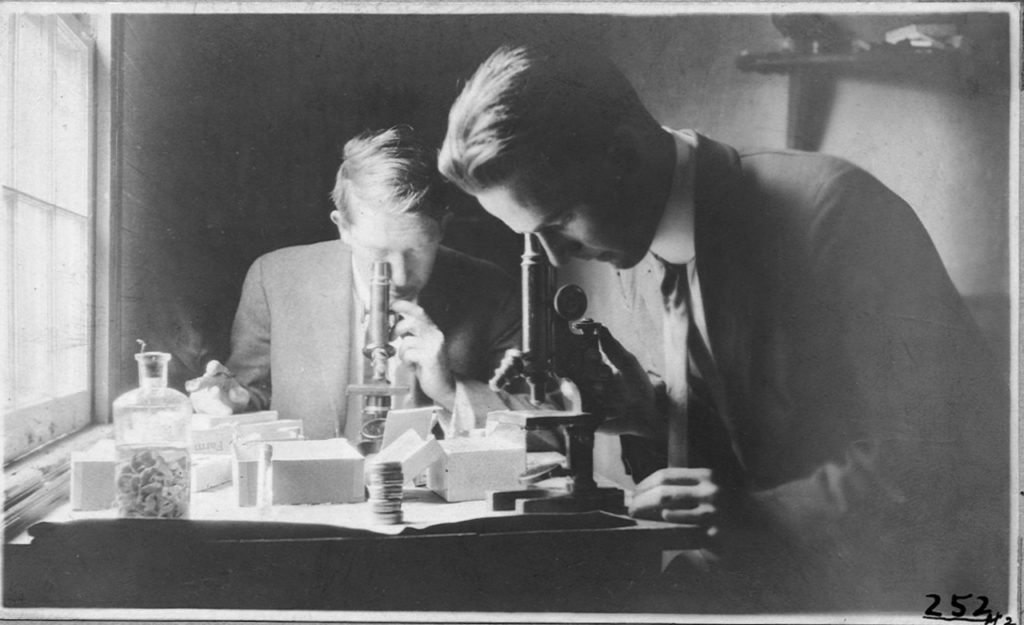 Black and white image of two scientist using microscopes surrounded by other scientific equipment.