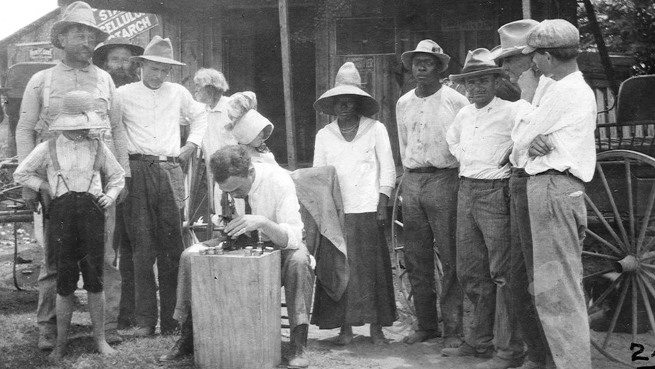 Black and white image of a group of men and children surrounding a scientist using a microscope in front of a store front.