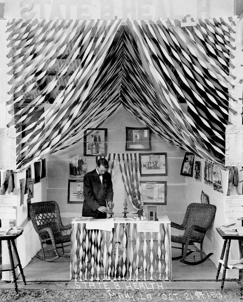 Black and white image of a State Board of health exhibition. Exhibition is lined with streamers from the sealing and front table. Exhibition has a small table with microscopes, chairs, and photographs.
