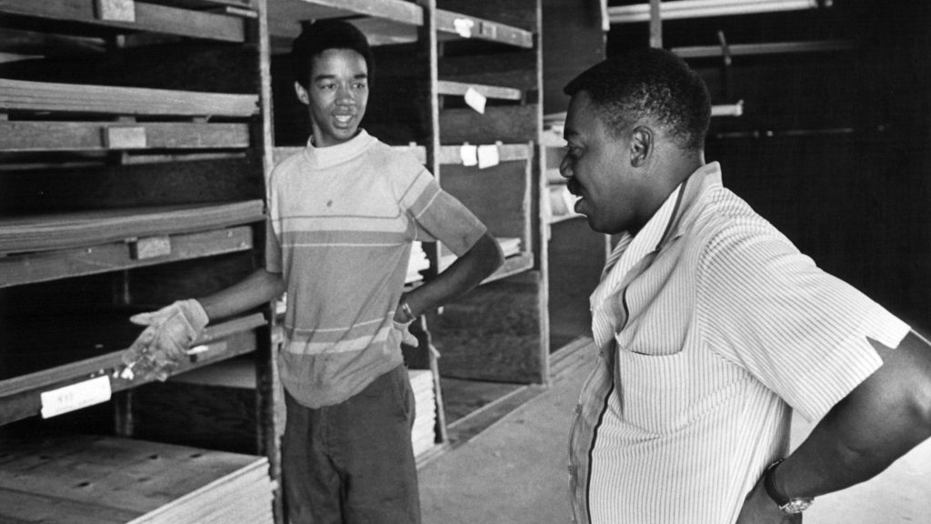 Black and white photo of two men discussing within a lumber yard in Detroit.