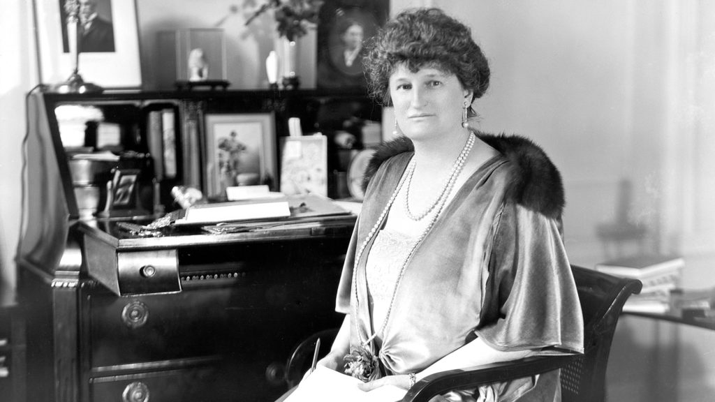 Black and white portrait of Abby Rockefeller. She is sitting in front of a large desk with other portraits.