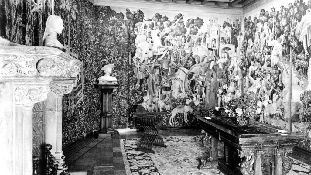 Another black and white image of a room in the Rockefeller home with a medieval stone fireplace and walls covered in unicorn tapestries.