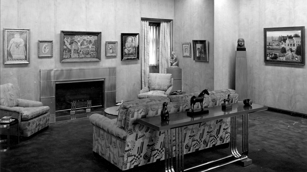 Black and white photos of a children’s playroom. Featured is a fire place with furniture, with the the walls lined with 7 hanging artworks, 2 statues and four small figurines.