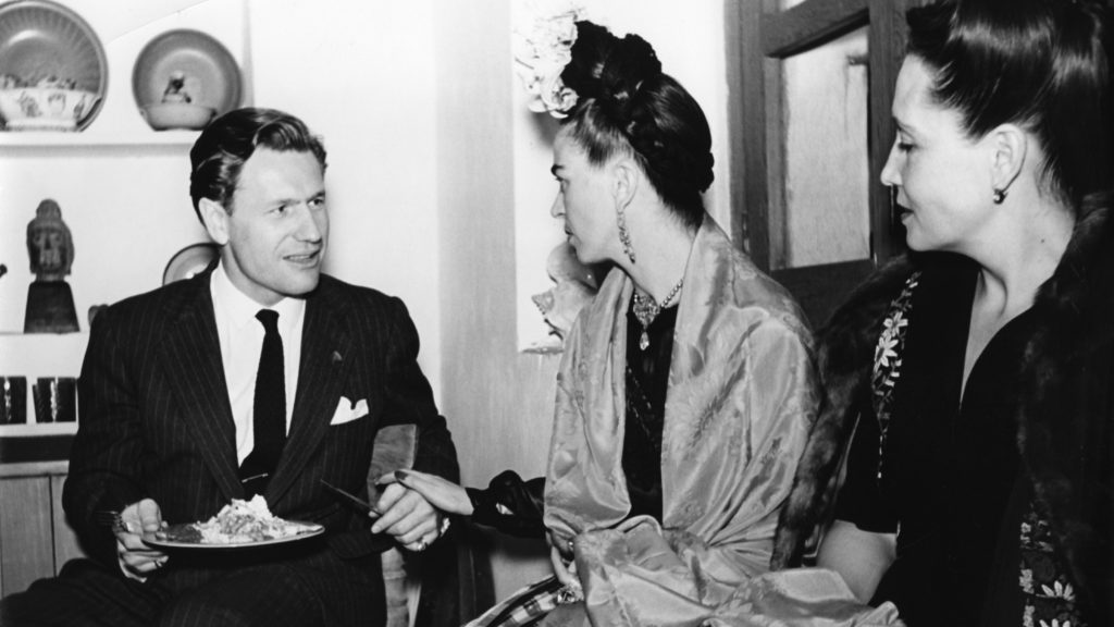 Black and white photo of Nelson Rockefeller and Frida Kahlo. They are seated to another unnamed woman. They are seated in front of artwork that lines the back wall.