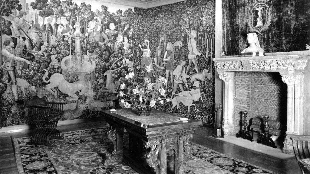 Black and white image of a room in the Rockefeller home with a medieval stone fireplace and walls covered in unicorn tapestries.