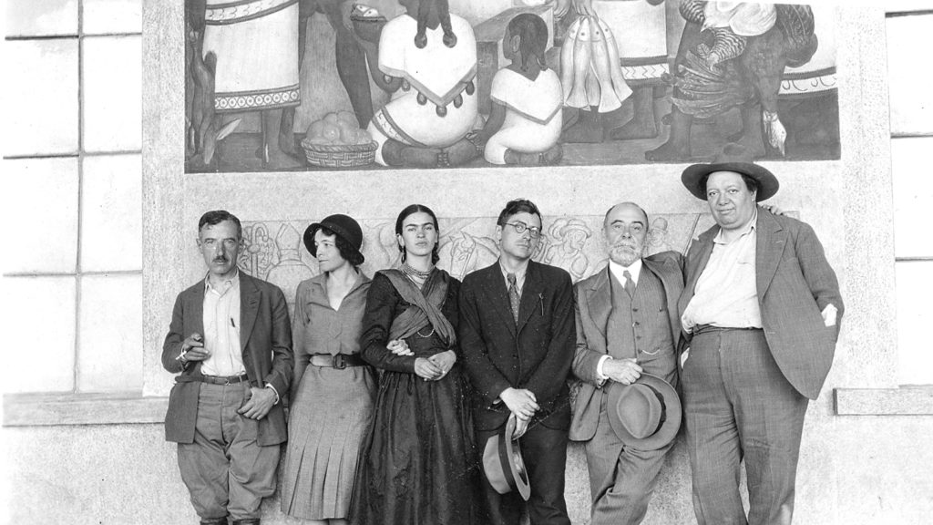Black and white photo of Frida Kahlo and Diego Riviera and several unnamed people. They are standing beneath Diego Riviera’s work.