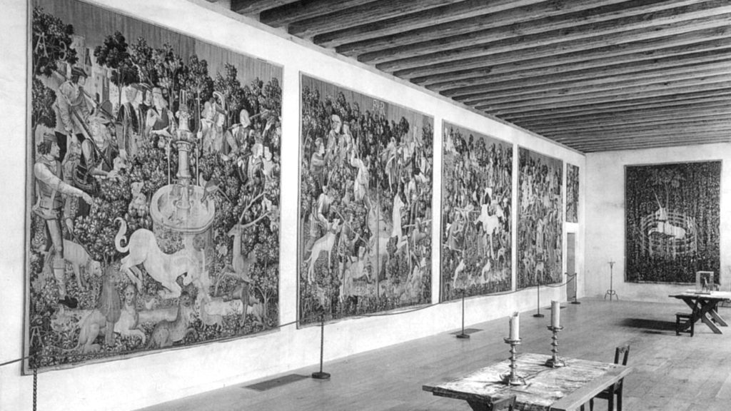 Large room at The Met Cloisters, with wooden ceiling and displaying four large medieval tapestries. The tapestries depict a large white unicorn being hunted by a large crowd of people.