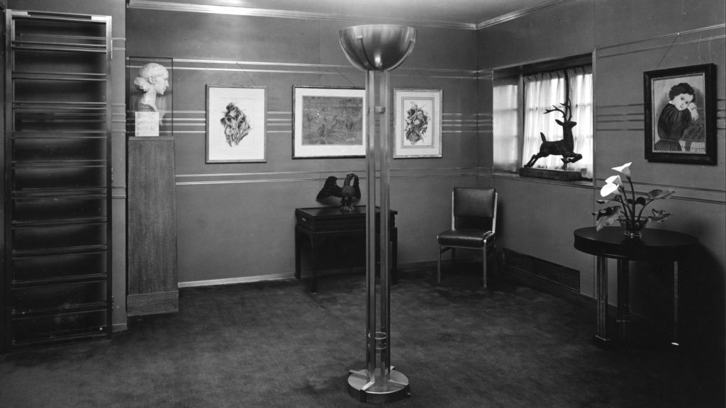 Black and white photo of a gallery within the residence with four hanging artworks, a chair, a large lamp, one head bust, and three figurines.