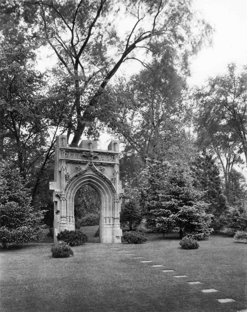 Medieval arch with a walkway leading to the arch in a black and white photo within a garden.