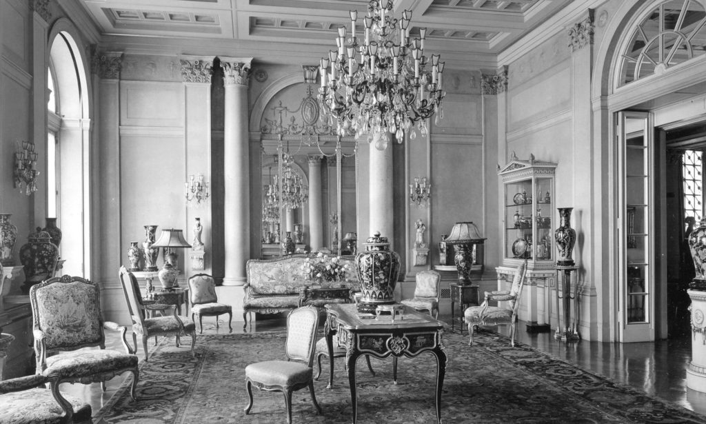 Black and white photo of the town house of Abby Rockefeller which features a large chandelier and cools lining the back wall. To the left is a large window and an entrance to another room in the Townhouse.