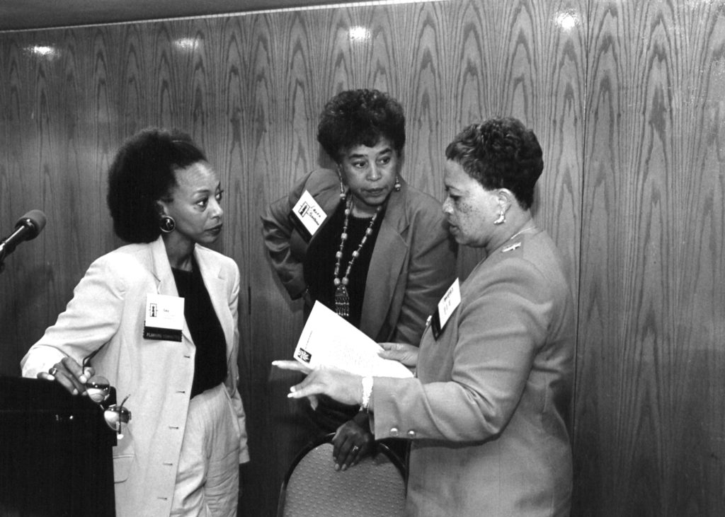 Three Black women lawyers from the Lawyers Committee for Civil Rights Under Law discuss together. One stands at a podium. 