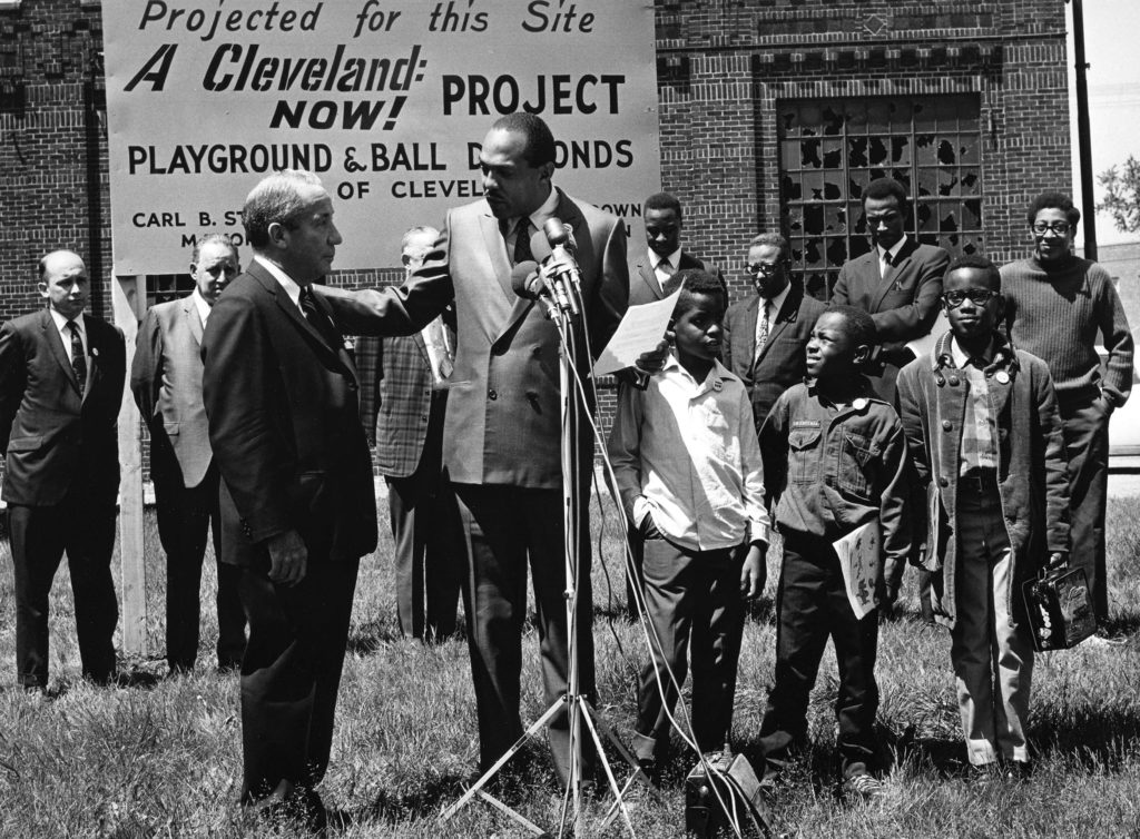 Cleveland Mayor Carl Stokes, the first Black mayor of Cleveland, stands at a microphone surrounded by Black and white men in suits and Black children. A sign reads "A Cleveland Now! Project. Playground and Ball Diamonds"