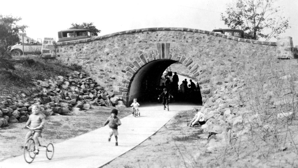 Black and white image of an underpass and bridge in Radburn, New Jersey. Cars are passing over the bridge, as children are running underneath. One child is riding a tricycle.
