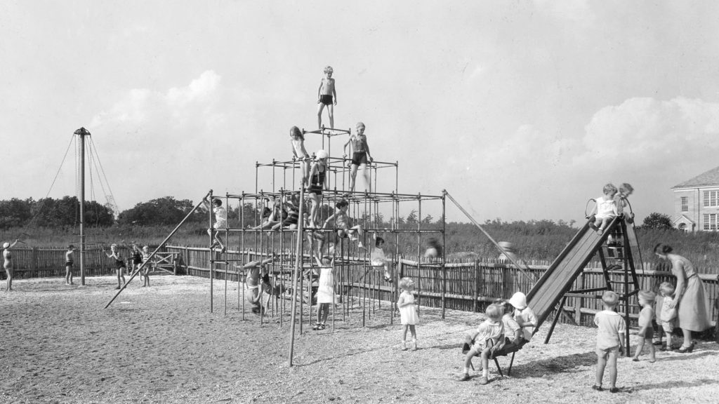 Black and white photo of children playing in a community playground. Featured is a large slide and play set within a fenced in area.