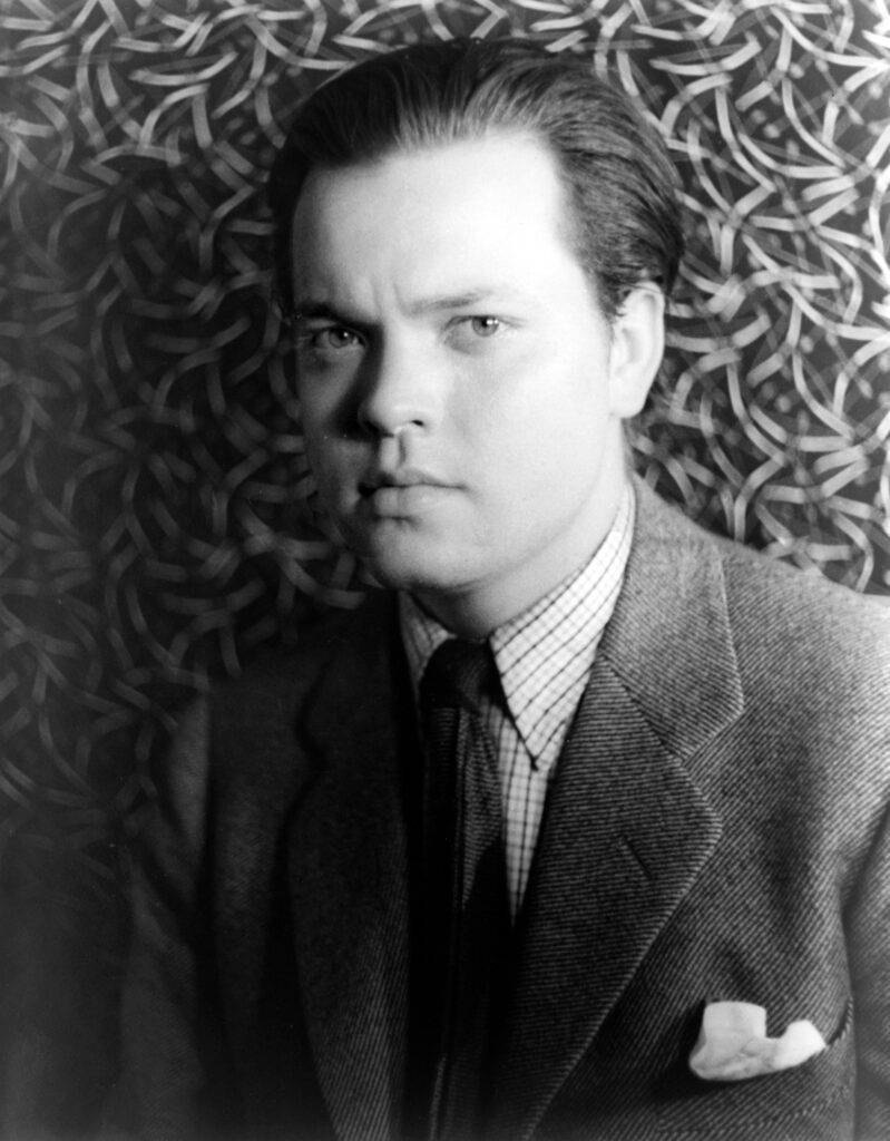 Black and white portrait of Orson Welles, the radio personality who performed "The War of the Worlds" in October 1938. Wells is wearing a suit and tie and looks sternly into the camera. The portrait is dated 1937.