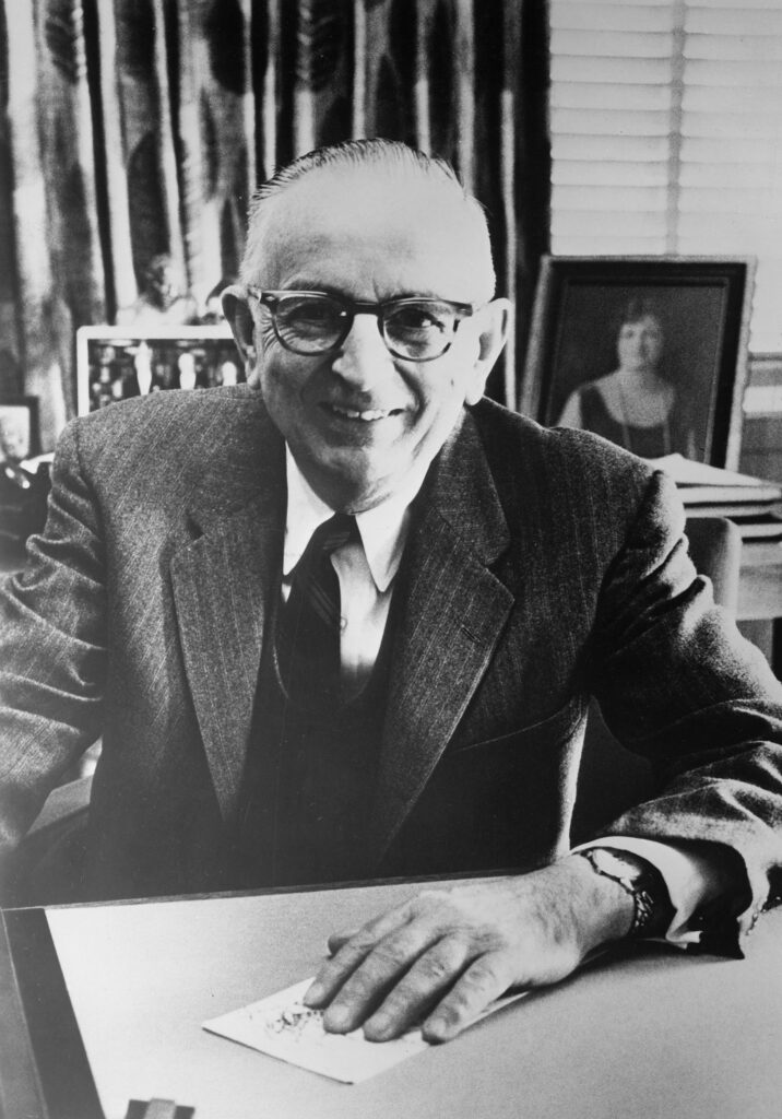 Portrait of Warren Weaver, Director of the Natural Sciences Division of the Rockefeller Foundation. Weaver is wearing glasses and is dressed in a suit and tie and sits, smiling, at a desk.