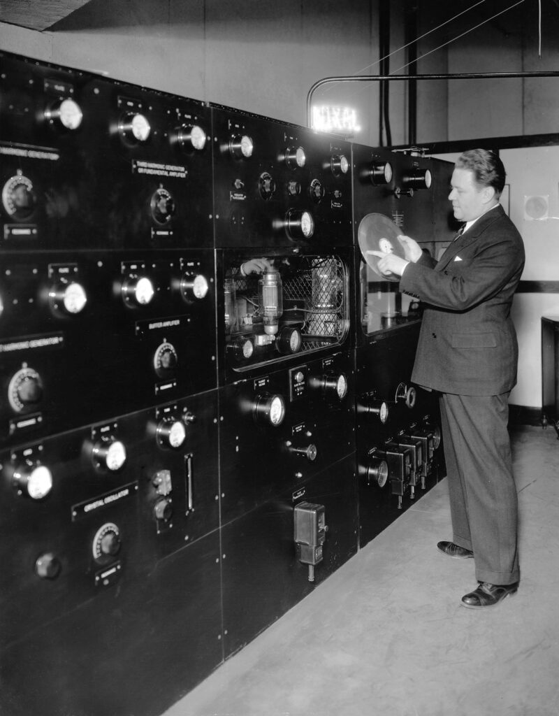 Black and white image shows a man examining a wall lined with radio broadcasting equipment, at radio station WIXAL in Boston, in 1937. The image is from the Rockefeller Foundation archival collections.