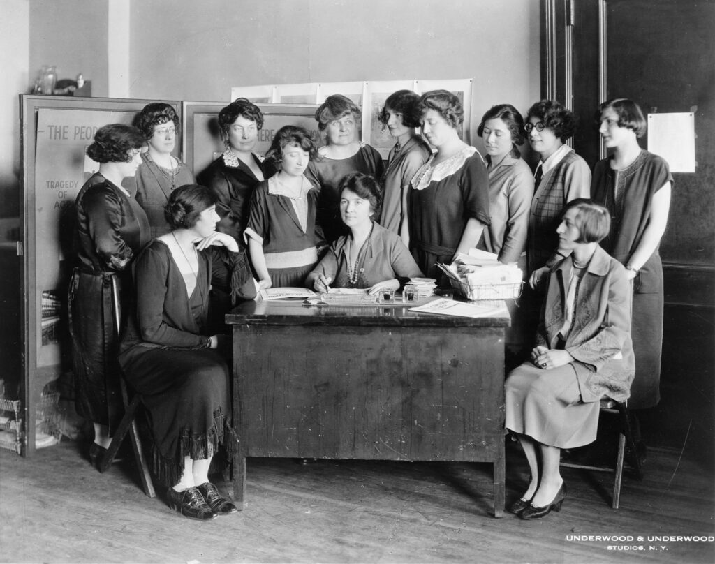 Black and white photograph of Margaret Sanger seated behind a desk with twelve other women surrounding her.