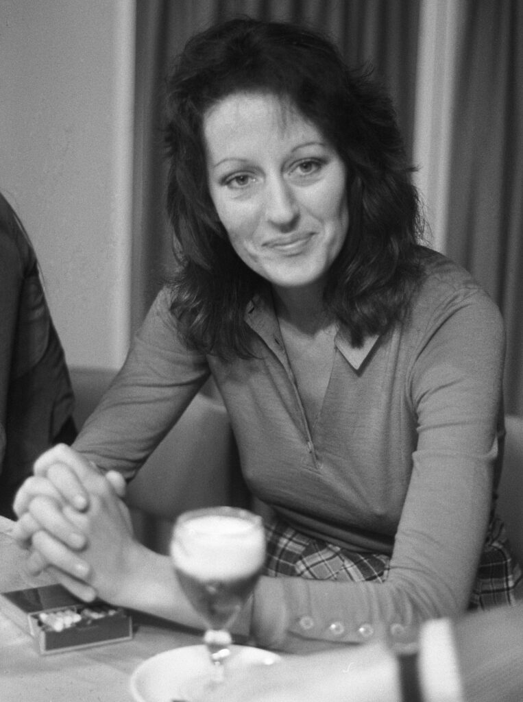 Germaine Greer sits at a table with her hands crossed, looking at the camera with a contented smile. A box of cigarettes and a glass of beer can be seen at her place setting on the table.