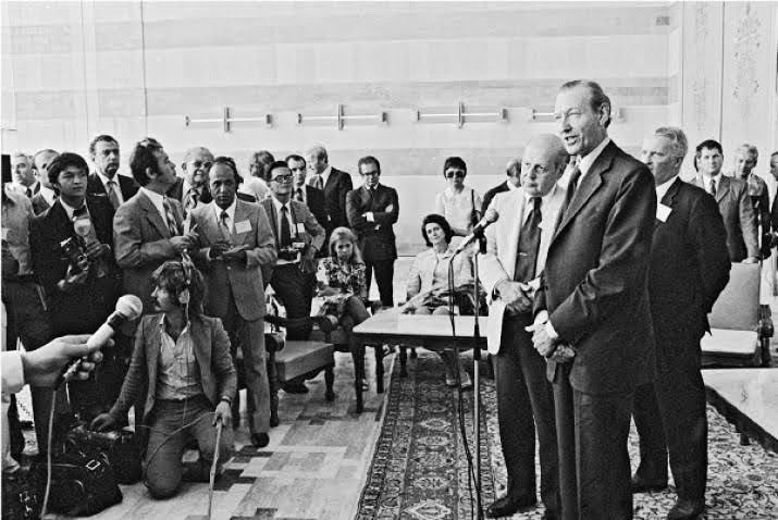 Two white men in suits and ties address a room full of mostly male journalists at the United Nations Population Conference in Bucharest in 1974.
